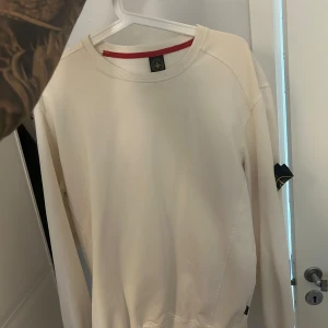 Vit sweatshirt från Stone Island - Säljer en clean vit sweatshirt från Stone Island med klassisk patch på vänster ärm. Tröjan har rund hals, långa ärmar och en röd detalj på insidan av kragen. Perfekt för dig som gillar stilrena och exklusiva plagg.