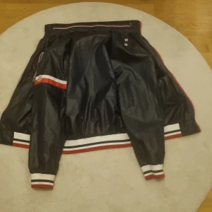 Moncler jacka i rött och svart - Snygg bomberjacka från Moncler med svart utsida och röd insida. Jackan har vita och röda detaljer, Moncler-logga på ärmen och ribbade muddar med ränder. Materialet är glansigt och känns sportigt, perfekt för dig som gillar streetstyle.