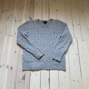 Grå kabelstickad tröja Polo Ralph Lauren - Kabelstickad grå tröja från Polo Ralph Lauren med klassisk rund hals och broderad vinröd logga på bröstet. Långärmad modell i mjuk bomull, perfekt för lager-på-lager och chill stil. Snygg ribbad kant vid ärmslut och nederkant. Storlek L 14/16