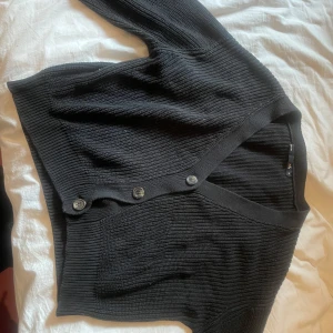 Svart ribbad kofta med knappar - Säljer en svart ribbad kofta från lager 157 i storlek S. Koftan har v-ringning, tre knappar framtill och är croppad i modellen. Perfekt att slänga över en topp eller klänning för en chill look.