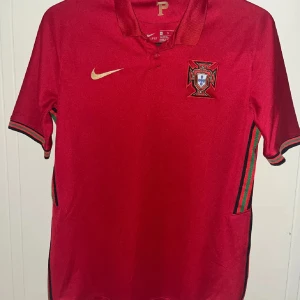 Portugal Ronaldo 7 Nike fotbollströja  - Snygg Portugal fotbollströja från Nike med Ronaldo 7 i guld på ryggen. Tröjan är röd med guldfärgade detaljer, har korta ärmar och krage med knapp. Broderat Portugal-emblem på bröstet och randiga gröna/röda detaljer på sidorna och ärmslut. Tillverkad i lätt polyester.