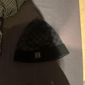 Svart Louis Vuitton mössa med rutmönster - Snygg svart mössa från Louis Vuitton med diskret rutmönster och klassisk LV-logga framtill på uppviket. Perfekt accessoar för dig som gillar exklusiv streetstyle och vill ha något som sticker ut. Passar både till jacka och hoodie.