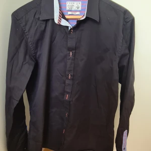 Svart skjorta från Jack & Jones L - Stilren svart skjorta från Jack & Jones i storlek L. Skjortan har långa ärmar, knappar framtill och coola detaljer med randigt och rutigt tyg på insidan av kragen och manschetterna. Perfekt för dig som vill ha en klassisk look med en twist.