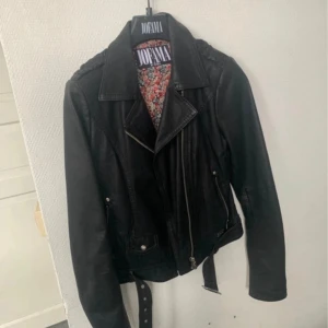 Svart skinnjacka Jofama by Kenza - Snygg svart skinnjacka från Jofama by Kenza med coola silvriga dragkedjor, bälte med nitar och blommigt foder. Klassisk bikerstil med bred krage och detaljer som ger jackan en edgy vibe. Perfekt för dig som vill sticka ut. Jackan är enbart använd ett par gånger och är i nyskick. Nypris 2999:-