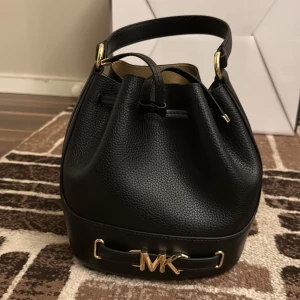 Svart bucket bag från Michael Kors - Snygg svart bucket bag från Michael Kors i äkta skinn med guldfärgade detaljer och MK-logo framtill. Väskan har snörning upptill, ett stilrent handtag och avtagbar axelrem. Insidan är beige och väskan har en klassisk, rundad form som passar till många outfits.