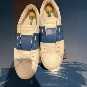 Valentino Garavani Open sneakers blå/vit - Äkta Snygga Valentino Garavani Open sneakers i vitt skinn med bred blå läderdetalj över vristen och ikoniska nitar på hälen. Kommer med box, skopåsar och oöppnade extra snören.  Klassisk rund tå, vita snören och platt sula. Bara att dra iväg ett meddelande ifall övriga frågor skulle uppstå. Priset är diskuterbart!