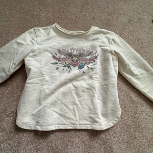 Vit långärmad tröja från Zadig & Voltaire - Vit långärmad tröja från Zadig & Voltaire i bomull. Framsidan har ett coolt tryck med blommor och text i rosa och blå toner. Tröjan har rund halsringning och avslappnad passform, perfekt för en chill och snygg look.