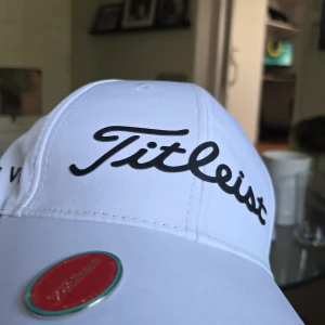Vit Titleist keps med logga - Snygg vit keps från Titleist med svart broderad logga framtill. Klassisk böjd skärm och justerbar passform. Perfekt för golfbanan eller som sportig accessoar. Tillverkad i lätt och bekvämt material.