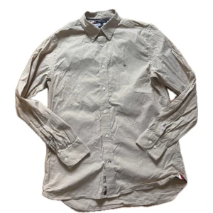 Beige skjorta Tommy Hilfiger M - Snygg beige skjorta från Tommy Hilfiger, modell THFlex Regular Fit. Klassisk krage, knappar hela vägen och diskret logga broderad på bröstet. Tillverkad i mjuk bomull och har långa ärmar med knapp vid manschetten. Perfekt för en clean och chill stil.