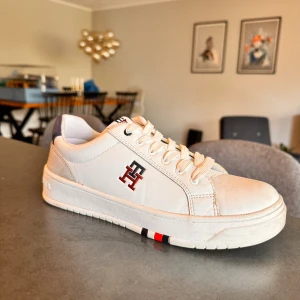 Vita sneakers från Tommy Hilfiger - Snygga vita sneakers från Tommy Hilfiger med klassisk logga på sidan och detaljer i rött, blått och vitt. Skorna har snörning, platt sula och är tillverkade i läder. Perfekta för en clean och stilren look.