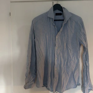 Ljusblå skjorta från Riley i bomull - Säljer en stilren ljusblå skjorta från Riley med klassisk krage och knäppning framtill. Skjortan är långärmad och tillverkad i mjuk bomull, perfekt för en clean och avslappnad look. Passar dig som gillar enkel och tidlös stil.