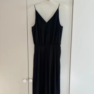 Svart v-ringad jumpsuit - Snygg svart jumpsuit från H&M med smala axelband och v-ringad hals. Jumpsuiten har ett brett midjeband. Materialet är mjukt och faller fint, perfekt för dig som gillar elegant look. Jumpsuiten är lite utsydd på sidorna