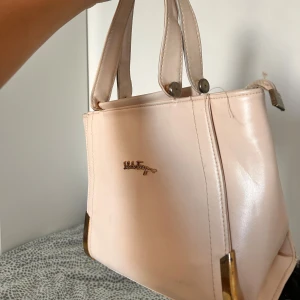 Ljusrosa handväska från Salvatore Ferragamo - Snygg ljusrosa handväska från Salvatore Ferragamo i skinn med guldiga metalldetaljer och logga framtill. Väskan har två handtag och ett rymligt innerfack med mönstrat foder och dragkedja. Perfekt för dig som vill ha en stilren och klassisk look.