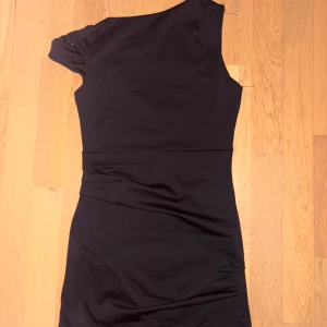 Svart oneshoulder kortklänning - Snygg svart kortklänning med oneshoulder-design och draperade detaljer vid midjan. Klänningen har en tight passform som framhäver figuren och är perfekt för dig som gillar stilrena och trendiga plagg. Materialet känns mjukt och följsamt mot kroppen.