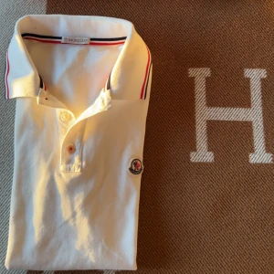 Vit Moncler pike - Vit Moncler pike  klassisk pikékrage och röd-blå randdetalj på kragen. Piken har korta ärmar, två knappar framtill och Moncler-logga broderad på bröstet. Materialet är mjuk bomull som känns skön mot huden. Perfekt för en clean och sportig look.