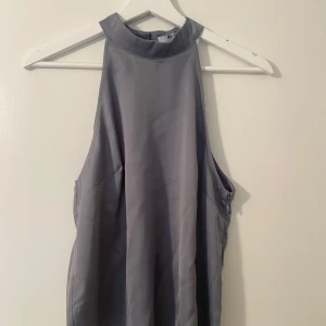 Grå ärmlös blus med hög krage - Snygg ”space grå” blus från NA-KD med hög krage och knäppning i nacken. Ärmlös modell med slät, mjuk yta och lätt glansigt tyg. Perfekt för dig som gillar stilrena och minimalistiska plagg.