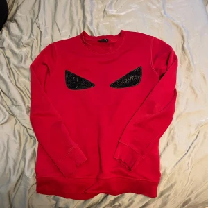 Röd Fendi sweatshirt med ögonmotiv - Cool röd sweatshirt från Fendi med svarta ögonmotiv framtill. Tröjan har rund hals, långa ärmar och är gjord i mjuk bomull. Perfekt statement piece för dig som vill sticka ut med en unik och edgy look. Storlek M alla spikar på bröstet sitter på endast slutet av armarna som det är en liten defekt men inget som märks av och knappt syns 