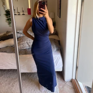 Marinblå långklänning med slits - Säljer en marinblå långklänning med ärmlös design och hög hals. Klänningen har en figurnära passform med snygg drapering i midjan och en diskret slits bak för extra rörelsefrihet. Perfekt för dig som vill ha en stilren och elegant look. Använd 1 gång på ett bröllop:)