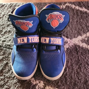 Adidas New York Knicks sneakers blå/orange - Unika Adidas sneakers i blått och orange med New York Knicks-logga på plösen och bred kardborre med texten 'New York'. Högt skaft, snörning och coola detaljer i svart och vitt. Perfekta för dig som älskar basket och streetstyle.