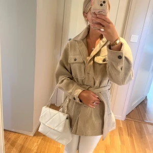 Beige overshirt med bälte och fickor - Snygg beige overshirt med knytskärp i midjan och stora bröstfickor med knappar. Jackan har en avslappnad passform och klassisk krage, perfekt att slänga över en hoodie eller t-shirt. Materialet känns mjukt och lite ulligt, vilket ger en chill men ändå uppklädd vibe.
