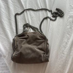 Grå axelväska Stella McCartney - Säljer en snygg grå axelväska från Stella McCartney med kedjeremmar i silver och coola detaljer längs kanterna. Väskan är gjord i mjuk mocka och har en rund logotypbricka. Perfekt för dig som vill ha en trendig och stilren väska. Den är äkta men har tyvärr inget äktahetsbevis på papper men har bild på att jag har köpt den i mina senaste beställningar. 