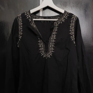 Svart blus med broderade detaljer - Svart blus med snygga vita broderier längs halsringningen, axlarna och ärmsluten. Långärmad modell med v-ringning och avslappnad passform. Perfekt för dig som gillar boho-vibbar och unika detaljer.