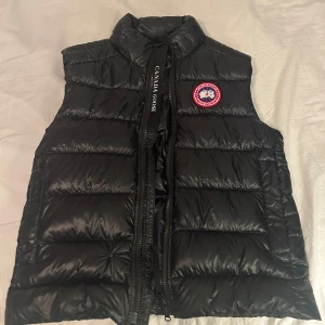Svart dunväst från Canada Goose - Snygg svart dunväst från Canada Goose med klassisk logga på bröstet. Västen är quiltad med breda kanaler, har hög krage och dragkedja framtill. Materialet är glansigt och känns riktigt lyxigt. Perfekt för lager-på-lager under kyliga dagar. Skicka vid frågor, kvitto finns också🙏