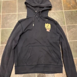 Svart hoodie från Morris med brodyr - Svart hoodie från Morris med guldfärgad brodyr och brittisk flagga på bröstet. Klassisk känguruficka, snörning i huvan och mjukt material. Perfekt för dig som gillar stilrena och snygga detaljer. Vid snabb affär kan ja sänka priset 