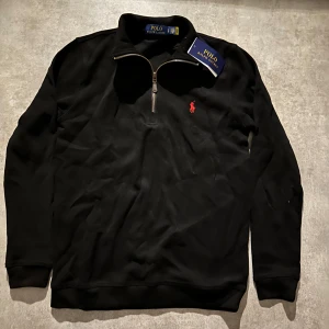 Svart half zip tröja Polo Ralph Lauren - Svart långärmad tröja från Polo Ralph Lauren med klassisk röd logga broderad på bröstet. Tröjan har en snygg half zip-dragkedja i metall och ribbade muddar. Perfekt för dig som gillar stilren och enkel design. Hör av vid minsta fundering 💫