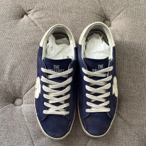 Blå sneakers från The Gallerist - Snygga blå sneakers i mocka från The Gallerist med vita detaljer och snörning. Skorna har en vit klöverdetalj på sidan och vit häl med logga. Kom privat för mer information.