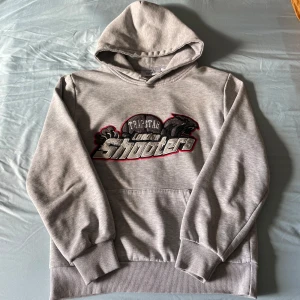 Trapstar London Shooters grå hoodie - Grå hoodie från Trapstar med broderad London Shooters-logga i svart, rött och silver på bröstet. Klassisk huva och stor magficka framtill. Mjuk bomullskänsla och ribbade muddar vid ärmslut och nederkant. Perfekt för dig som gillar streetwear.