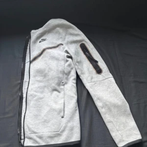 Grå Nike jacka med dragkedja - Snygg grå jacka från Nike med svart logga på bröstet och coola dragkedjedetaljer, bland annat på ärmen. Jackan har lång ärm och är tillverkad i mjukt bomullsmaterial. Perfekt för dig som gillar sportig och clean stil.