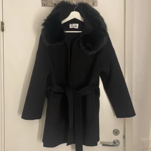 Svart kappa med fuskpäls från FUR Stockholm - Snygg svart kappa från FUR Stockholm med stor huva och bred krage med päls. Kappan har knytskärp i midjan och två stora fickor framtill. Perfekt för kalla dagar när du vill vara både varm och stilren. (Använd 1 gång)