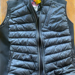 Svart dunväst från Parajumpers M - Snygg svart dunväst från Parajumpers i storlek M. Västen har quiltad design, dragkedja framtill och en bröstficka med dragkedja. Perfekt för lager-på-lager och har en sportig vibe med diskreta detaljer och klassisk look. Det ända negativa är det lilla hålet som syns på bilden men syns knappast!