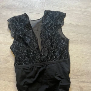 Svart spetsbody med blommönster - Snygg svart body med transparent mesh upptill och detaljerad spets med blommigt mönster. Djup v-ringning framtill och bak. Pieces, storlek xs
