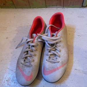 Nike fotbollsskor i grått och rött - Säljer ett par Nike fotbollsskor i grått med rosa detaljer och innersida. Skorna har snörning och dobbar för bra grepp på planen. Swoosh-logga på sidan och undersidan. Perfekta för dig som vill sticka ut på matchen!