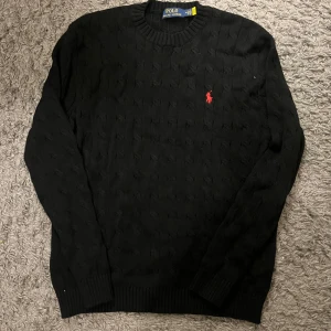 Svart kabelstickad tröja Ralph Lauren - Svart kabelstickad tröja från Polo Ralph Lauren med klassisk röd broderad logga på bröstet. Tröjan har rund halsringning, ribbade muddar och lång ärm. Perfekt för dig som gillar stilren och tidlös design.