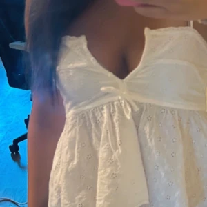 Vit somrig topp med rosett - Supersöt vit topp  med broderade detaljer och liten rosett framtill. Klänningen har smala axelband och v-ringning, perfekt för varma dagar. Lätt och luftig modell som ger en romantisk känsla.