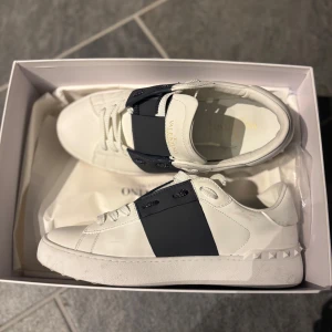Valentino Garavani Open Sneakers vit/blå - Snygga Valentino Garavani Open Sneakers i vitt kalvskinn med bred marinblå rem över vristen och ikoniska nitar på hälen. Klassisk rund tå, platt sula och vita snören. Perfekta för dig som vill ha en clean och lyxig look.