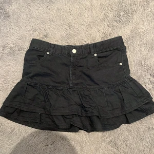 Svart volangkjol från Zara - Supersnygg svart kortkjol från Zara med volangkant och klassisk jeanslook. Kjolen har fickor fram, bälteshällor och stängs med knapp och dragkedja. Perfekt för dig som gillar en trendig och cool stil.