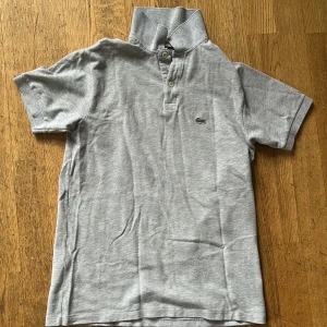 Grå Lacoste piké t-shirt - Snygg grå piké från Lacoste med klassisk krage och två knappar framtill. Ikonisk broderad krokodil på bröstet. T-shirten är kortärmad och gjord i mjuk bomull som är skön mot huden. Perfekt för dig som gillar stilren och sportig vibe.