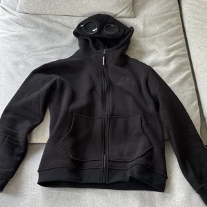 CP company hoodie - Svart hoodiejacka från C.P. Company med ikoniska goggles i huvan. Jackan har dragkedja framtill, två stora fickor och broderad logga på bröstet. Tillverkad i mjuk bomull och perfekt för dig som vill sticka ut med streetwear-vibe. Nyskick, passar även S bra.