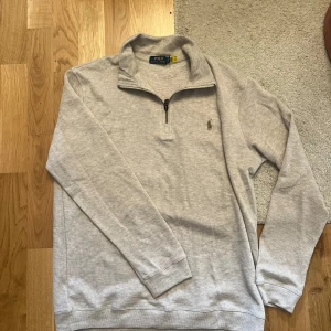 Polo Ralph Lauren Halfzip - Snygg ljusgrå långärmad tröja från Polo Ralph Lauren med klassisk half zip-dragkedja och broderad logga på bröstet. Mjuk bomullskvalitet och normal passform, perfekt för en clean och stilren look.