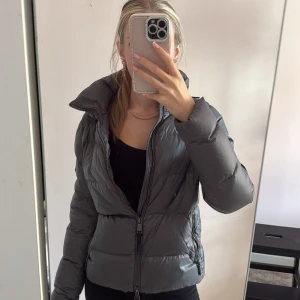 Grå pufferjacka från Polo Ralph Lauren - Snygg grå pufferjacka från Polo Ralph Lauren med hög krage och dragkedja framtill. Jackan har quiltad design, två sidofickor med dragkedja och Polo-logga på ärmen. Perfekt för kyliga dagar och riktigt skön att bära. Små små skador efter att den används