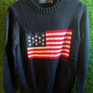 Mörkblå stickad tröja Polo Ralph Lauren - Snygg mörkblå stickad tröja från Polo Ralph Lauren med amerikansk flagga i rött, vitt och blått på bröstet samt RL-broderi. Rund hals och långa ärmar med ribbade muddar. Perfekt för dig som gillar klassisk preppy stil och vill sticka ut.