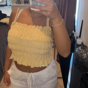 Gul croppad linnetopp med volang - Superfin ljusgul croppad linnetopp med volangkant och smockad struktur. Toppen är ärmlös med smala axelband och har en söt, somrig vibe. Perfekt till varma dagar och enkel att matcha med shorts eller kjol.