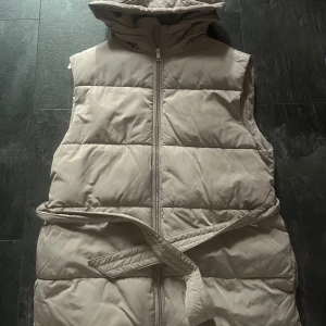 Beige quiltad dunväst från Nelly - Säljer en beige quiltad dunväst från Nelly i storlek 36. Västen har huva, dragkedja framtill och ett bälte i midjan för extra snygg passform. Perfekt att slänga över en hoodie eller stickad tröja när det är kyligt ute.