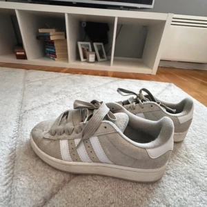 Adidas Campus i beige  - Säljer ett par Adidas Campus sneakers i beige  mocka med klassiska vita ränder på sidorna. Skorna har rund tå, platt sula och breda snören. Perfekta för dig som gillar en clean och stilren look.