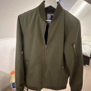 Grön  bomberjacka Jack & Jones - Snygg olivgrön bomberjacka från Jack & Jones. Jackan har dragkedja framtill, två fickor med dragkedja och en extra ficka på ärmen. Kanppt använd och i fint skick. Priset går alltid att diskutera. Skriv gärna för minsta intresse 
