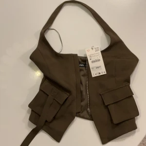 Olivgrön korsettopp från Zara med fickor - Snygg korsettopp från Zara med breda axelband och coola fickdetaljer framtill. Toppen har dragkedja framtill och en unik formad nederkant. Perfekt för dig som vill sticka ut med en trendig och edgy look. Plagget har aldrig använts.